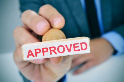 a young caucasian man at his office desk with a rubber stamp with the word approved